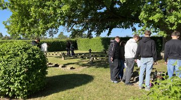 Auf dem Bild sind Schülerinnen und Schüler auf der KRiegsgräberstätte zu sehen. Sie stehen in kleinen Gruppen zusammen und lesen die Inschriften auf den Gräbern und auf einem Gedenkstein.